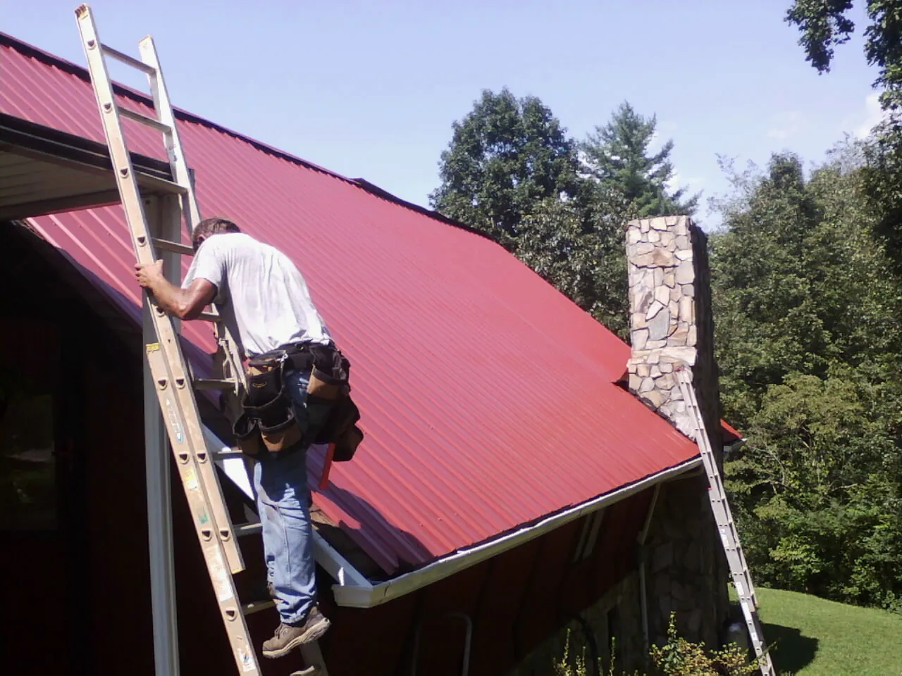 Roofer on a ladder beside a red metal roof and stone chimney — Soffit & Fascia Repair by Roofing Brothers in Elora